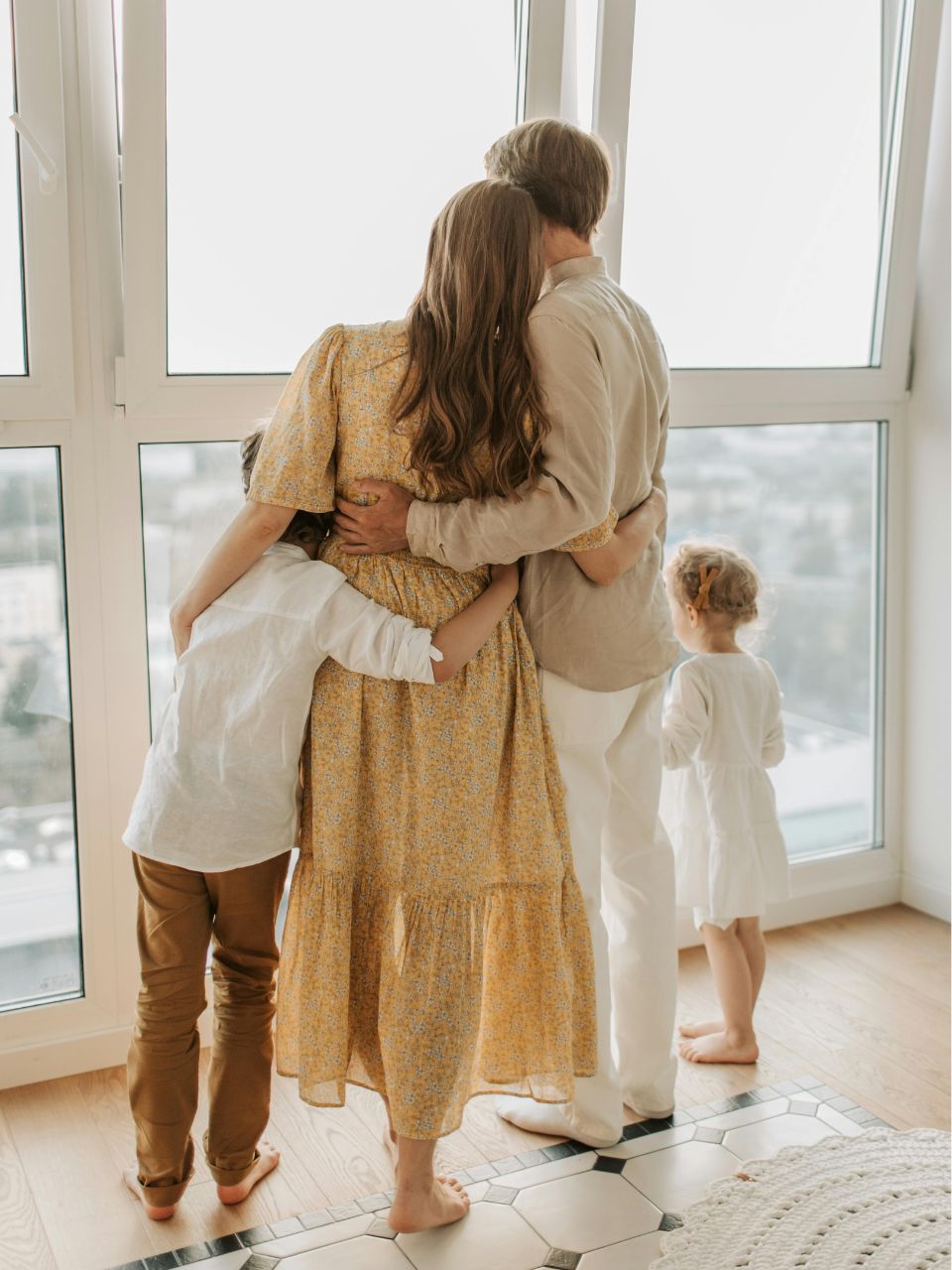 Family embracing near a window. Two children hug their parents. Soft colors, neutral setting.