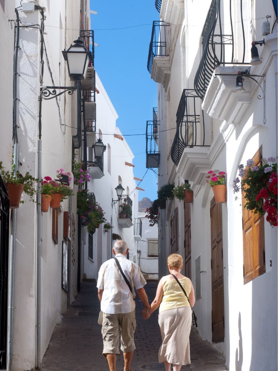 Couple holding hands walks down a narrow, white-walled street lined with flowerpots.