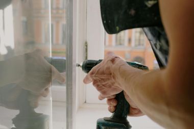 Hand drilling into a white window frame beside a reflective glass pane
