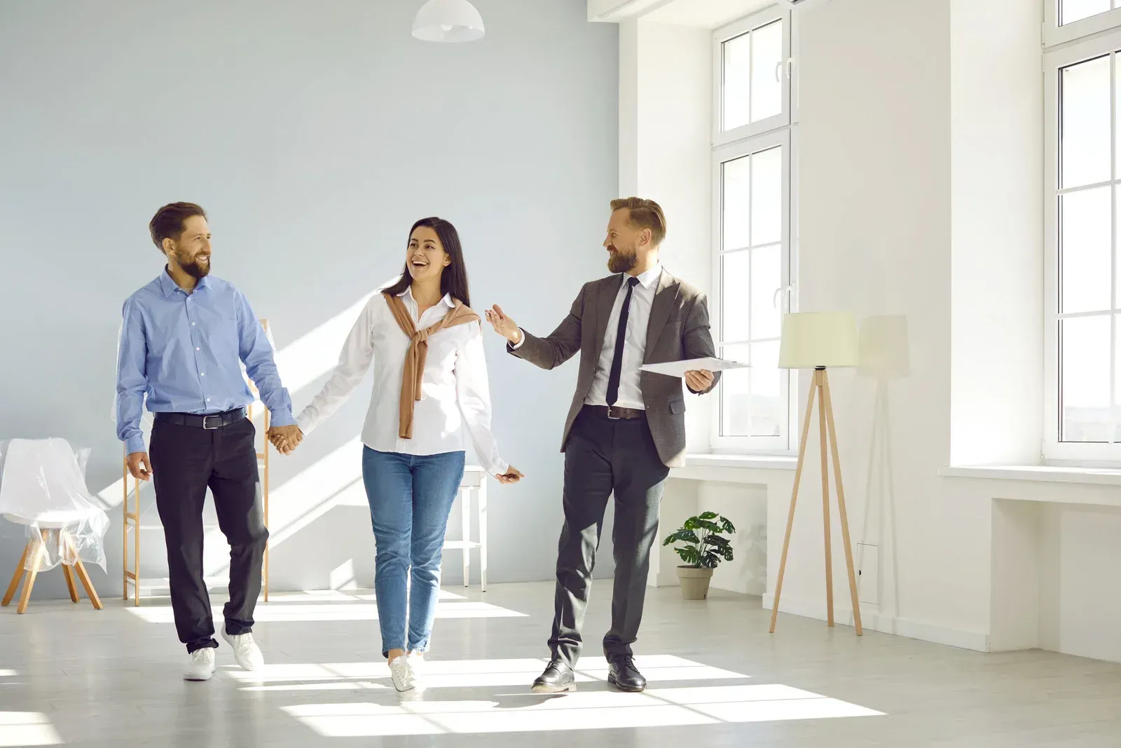 A real estate agent shows a couple an empty, light-filled room. The couple holds hands and smiles.