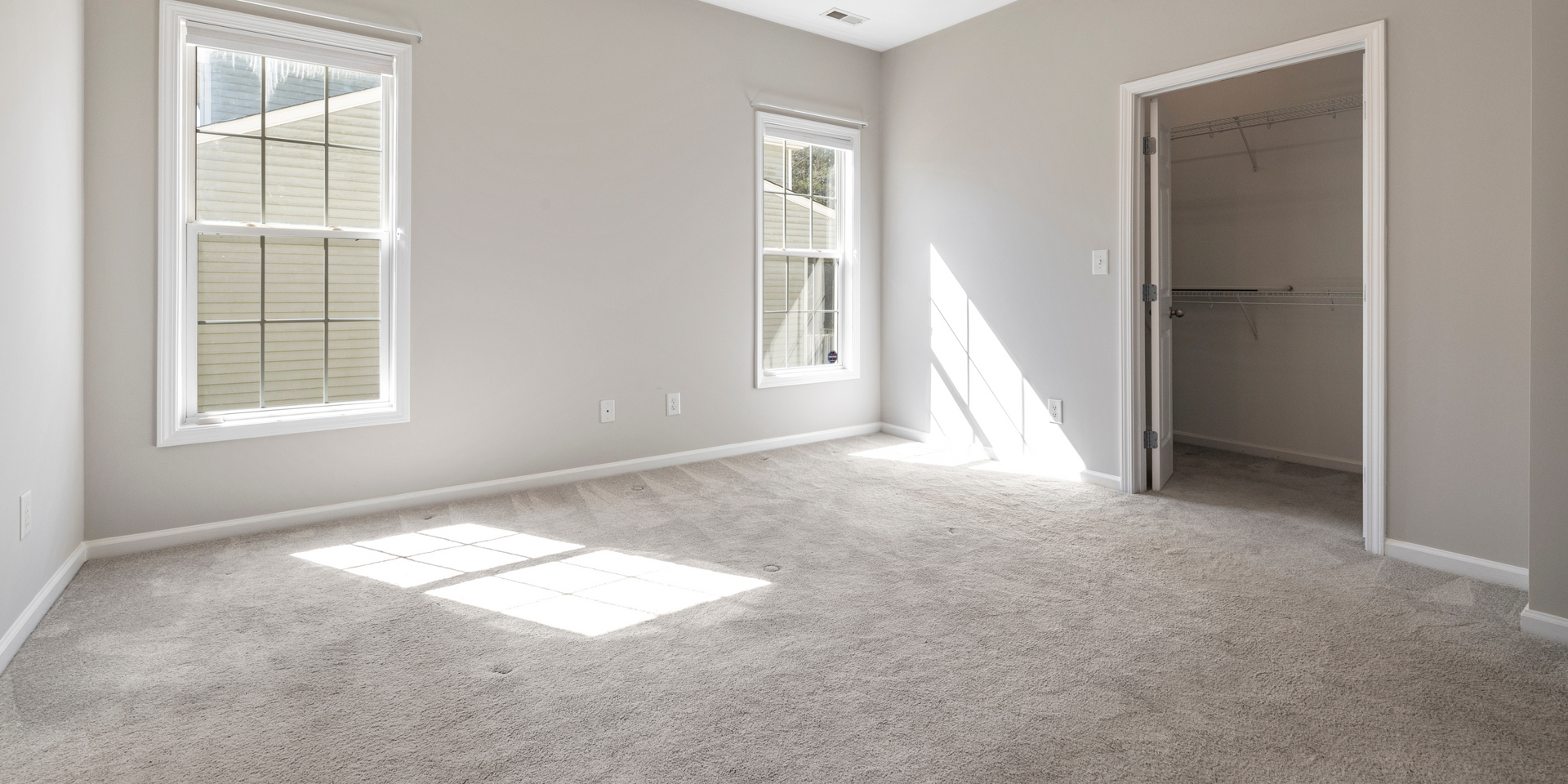 Empty bedroom with two windows, a closet, and neutral-colored carpet. Sunlight streams through the windows.