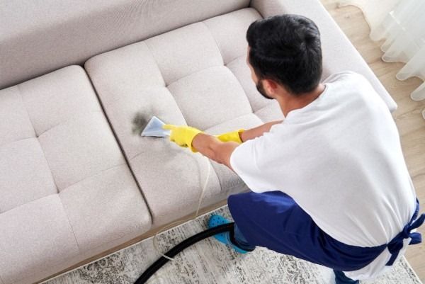 Man in gloves cleaning a beige sofa with a machine, indoors.