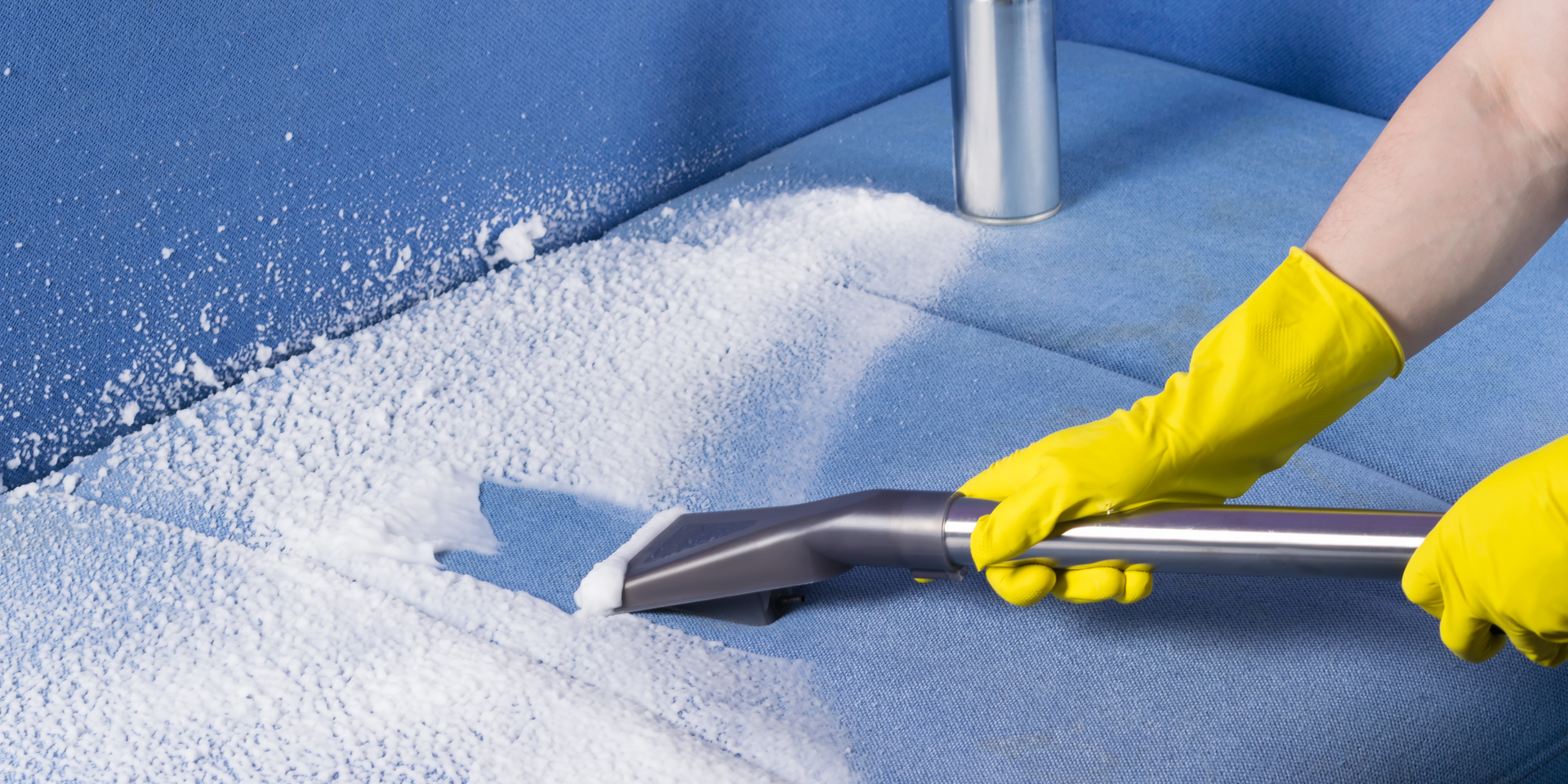 Person wearing yellow gloves cleaning a blue couch with a vacuum cleaner.