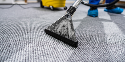 A carpet cleaner's nozzle is in use on a gray carpet. In the background, a yellow vacuum cleaner and blue shoe covers.