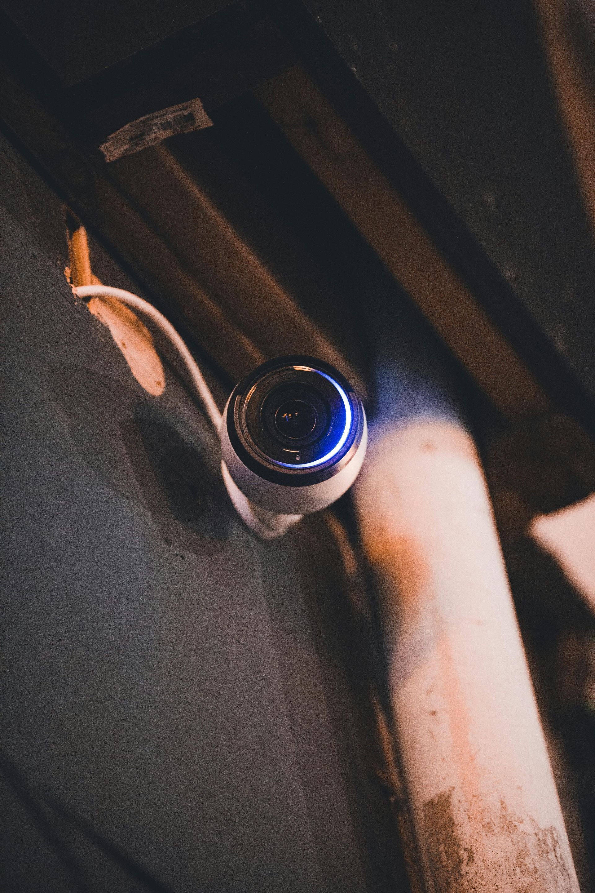 A small, white security camera with a blue ring, mounted on a dark wall near a pipe.