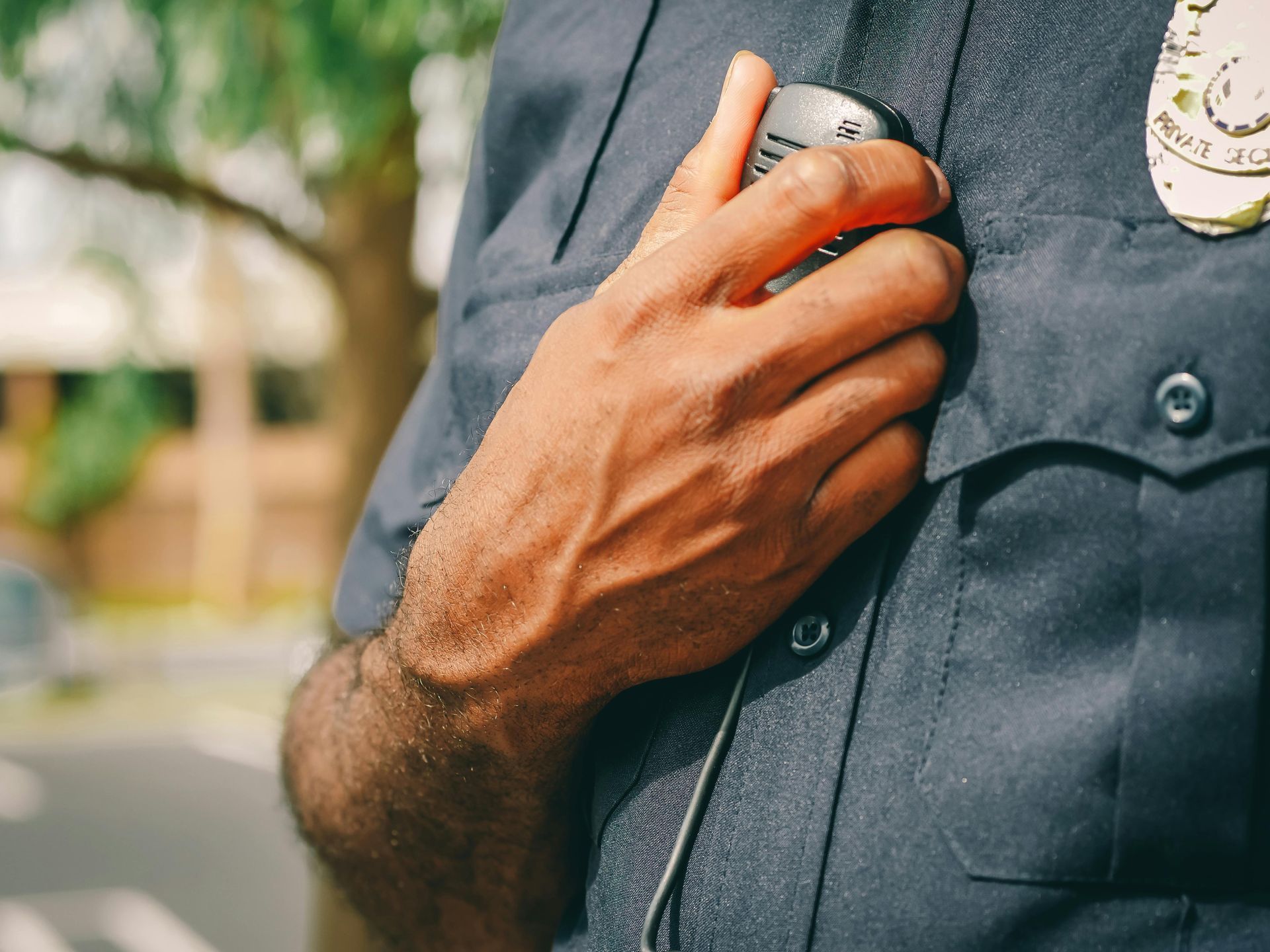 Close-up of a police officer's hand holding a radio microphone on their uniform, outdoors. The officer's skin tone is dark; blue uniform.