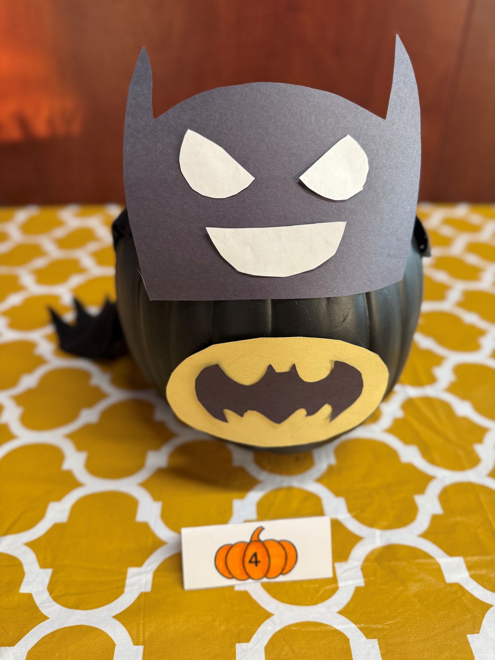 Batman pumpkin with paper mask and emblem, sitting on a yellow patterned table.