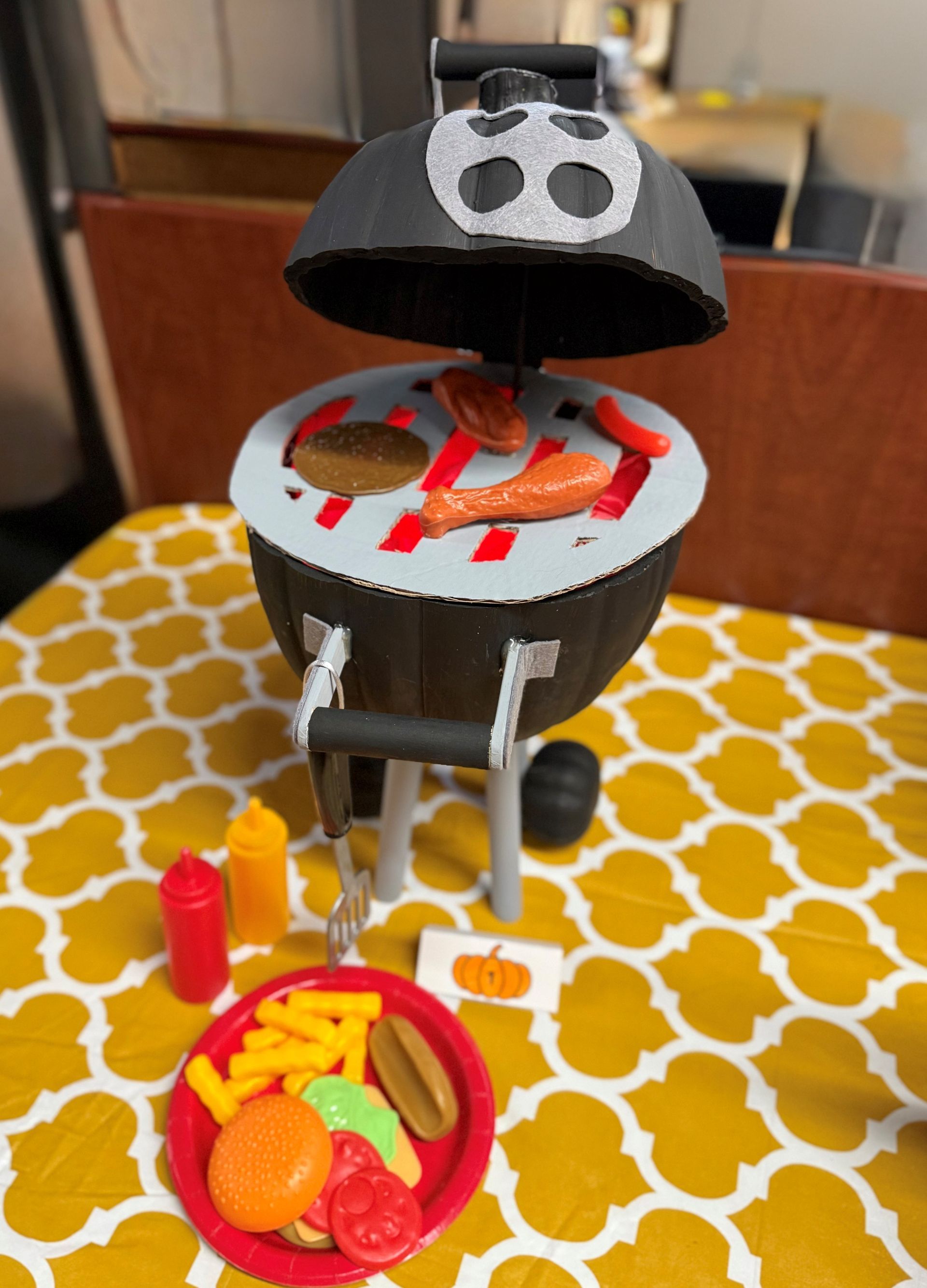 Toy grill with food, condiments, and a plate of food on a yellow patterned surface.
