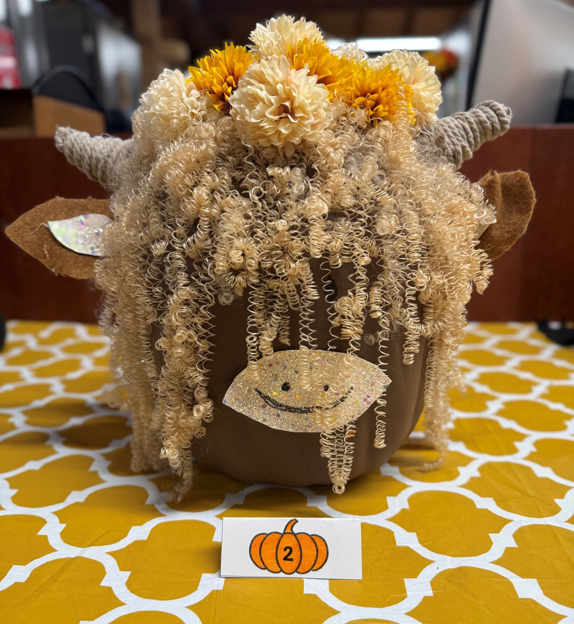 Pumpkin decorated as a Highland cow with tan and brown fur, horns, and a floral crown, on a patterned tablecloth.