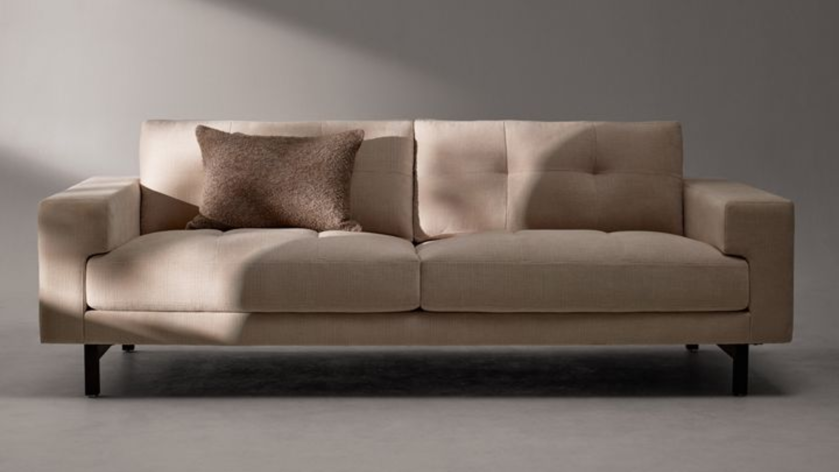 Beige two-seater sofa with a brown pillow on a light gray background; natural light.