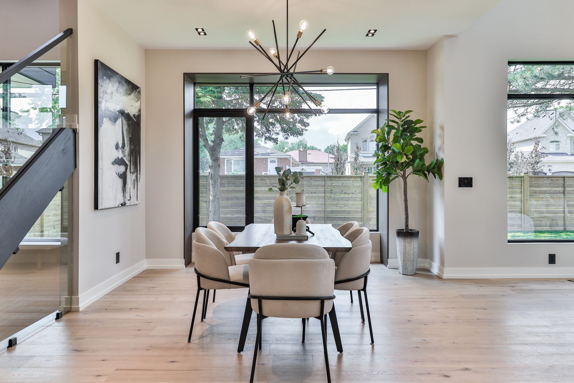 Dining room with light wood floors, modern chairs, table, and a sputnik chandelier. Large windows reveal a backyard and a black and white portrait.