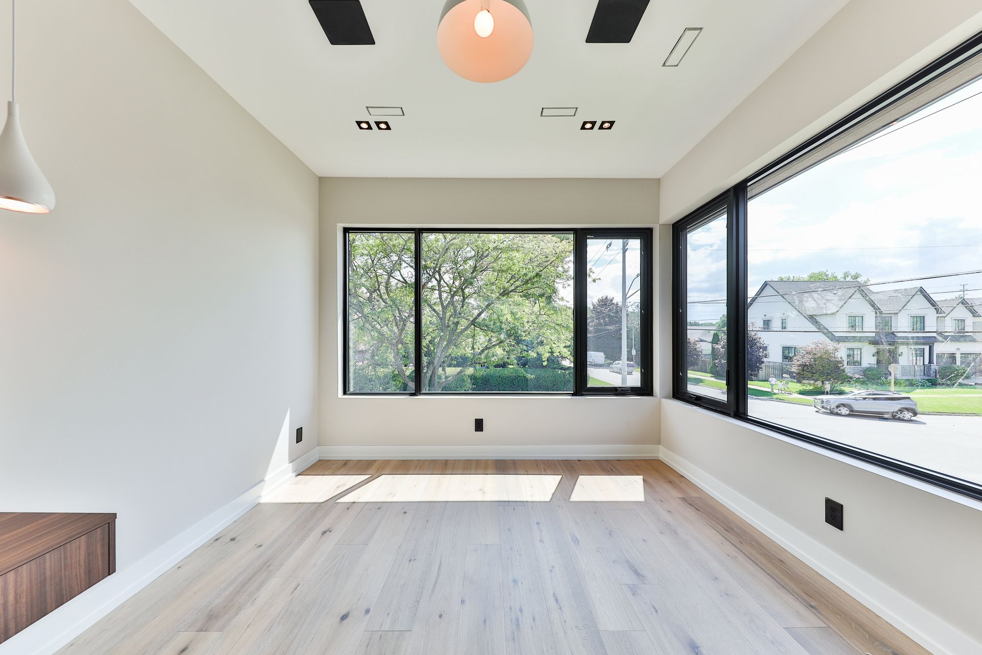Empty room with large windows offering a view of houses and trees. Neutral walls, light wood floor, modern lighting.