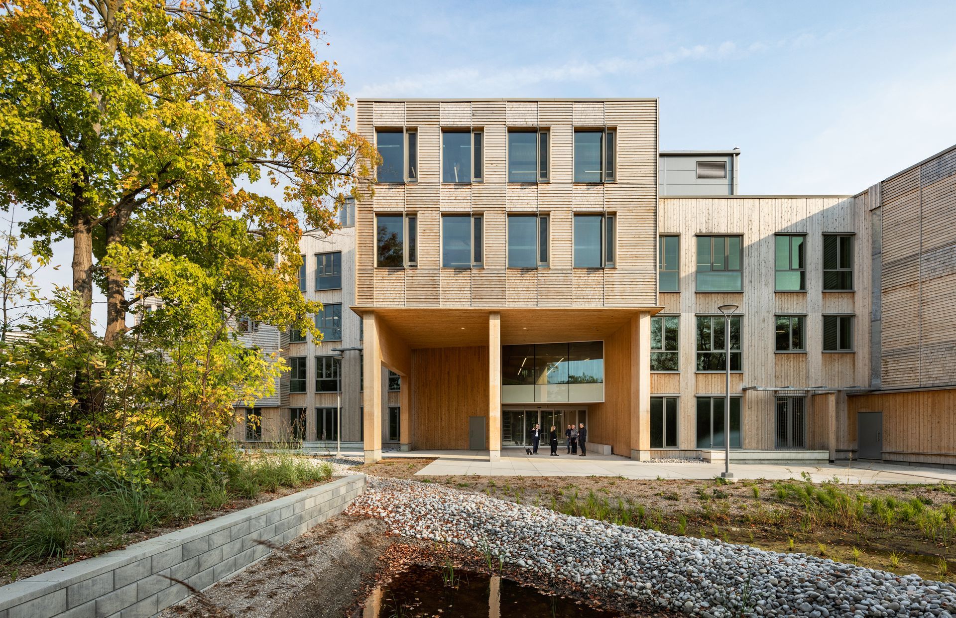 Wooden building with large open entrance, surrounded by trees and a rock bed. People can be seen inside the entrance.