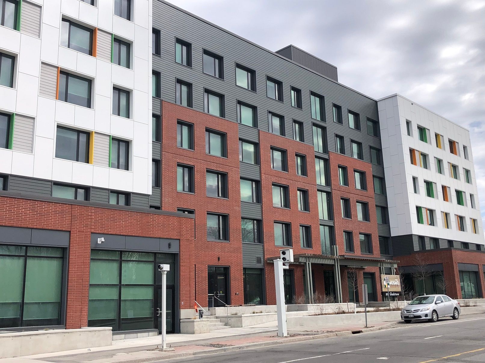Modern multi-story building with brick, white, and gray facades and large windows. A car and construction equipment are visible.