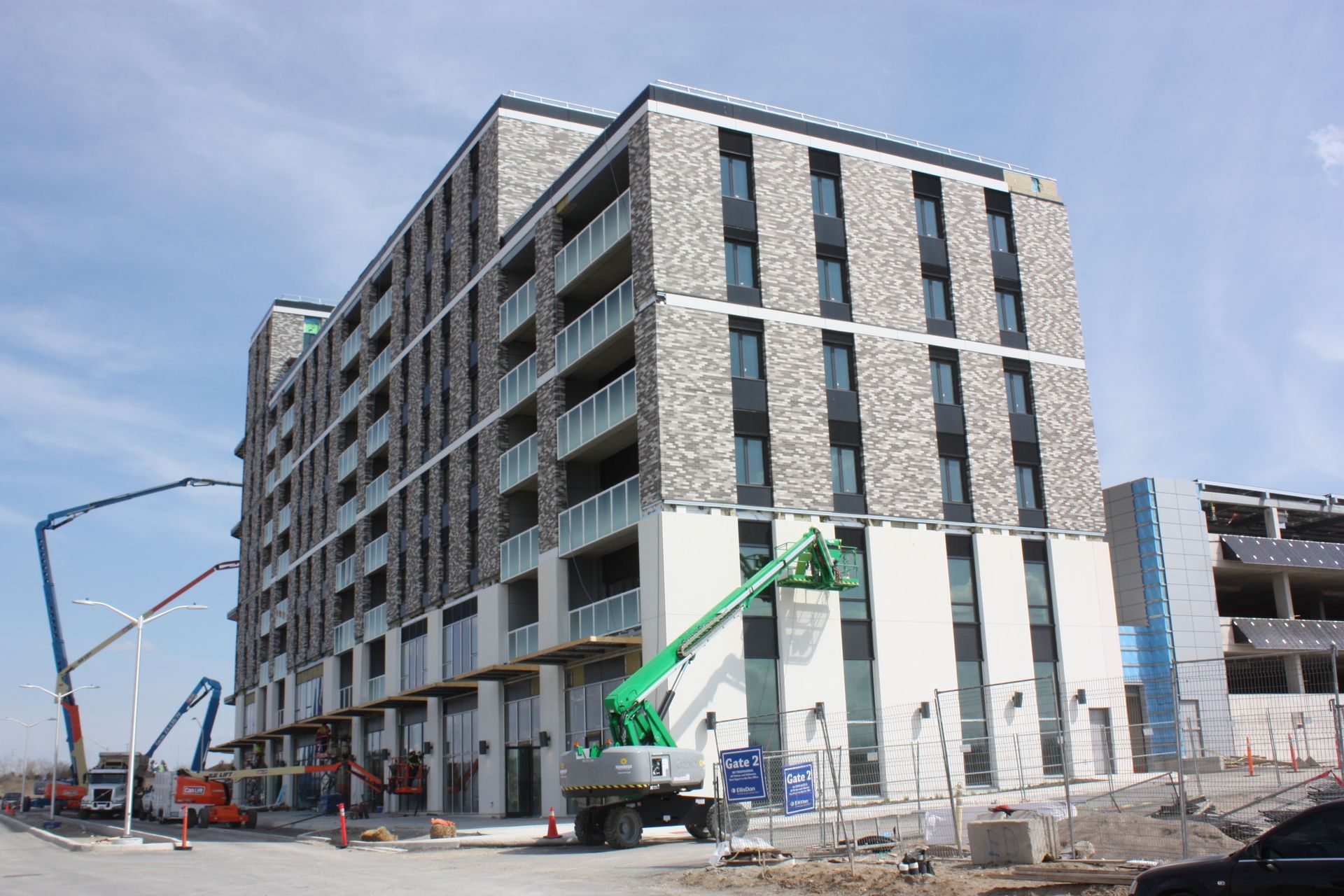 A multi-story apartment building under construction with a boom lift, cement trucks, and clear sky. The lower levels have white facades.