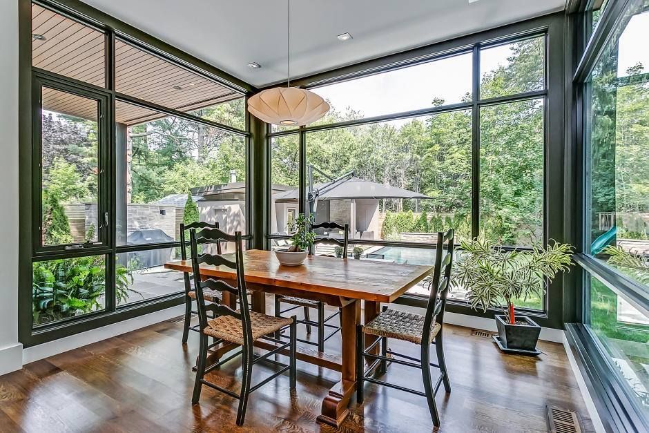 A dining room with large windows offering a view of lush greenery, featuring a wooden table and chairs, with natural light illuminating the interior.