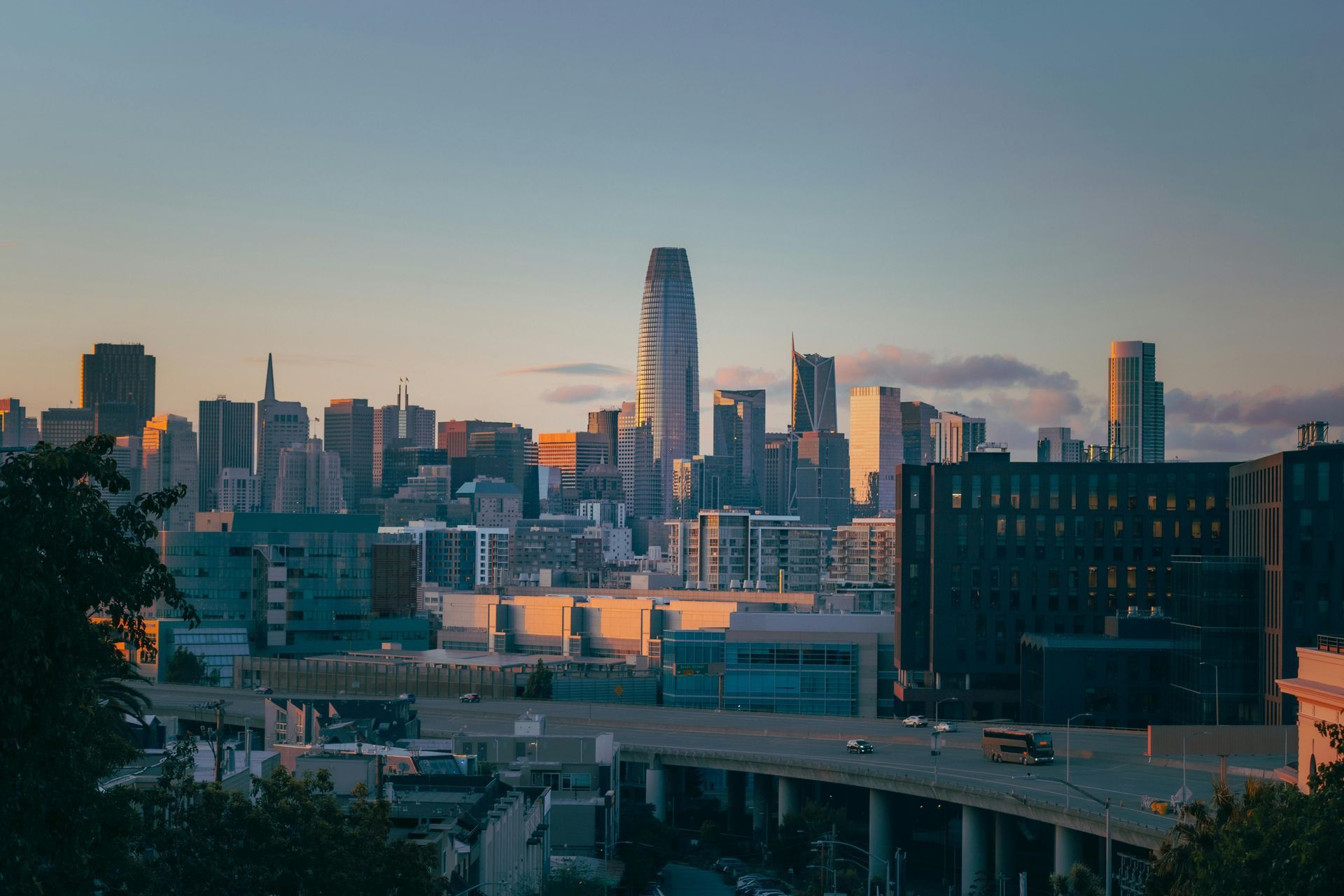City skyline at sunset with tall buildings, highway, and bridge; gold and blue hues.
