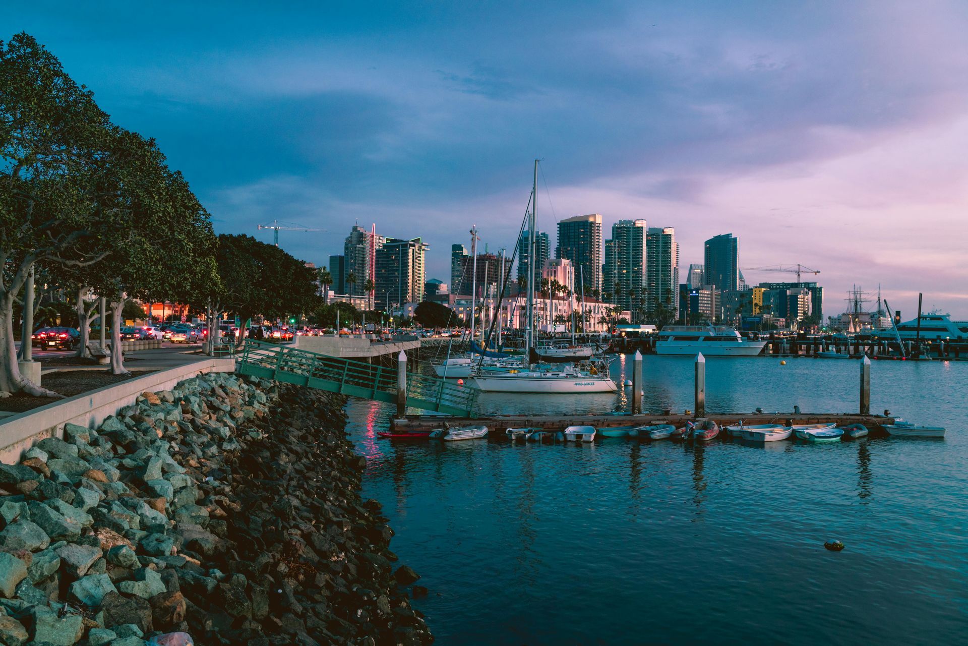Waterfront view of a city skyline at dusk; boats docked at pier, trees and pedestrians line the shore.