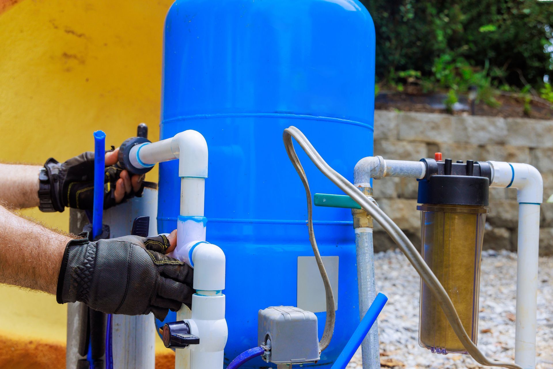 Hands with gloves connecting white PVC pipes to a blue water tank and filter outside.