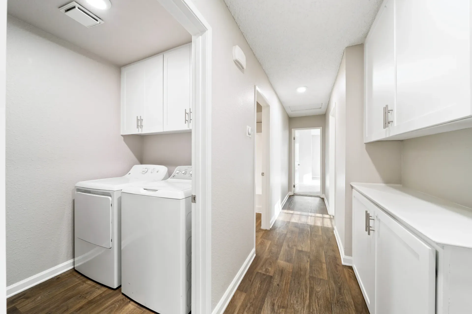 Laundry closet with a white washer and dryer, white cabinets, and a hallway with wood-look flooring.