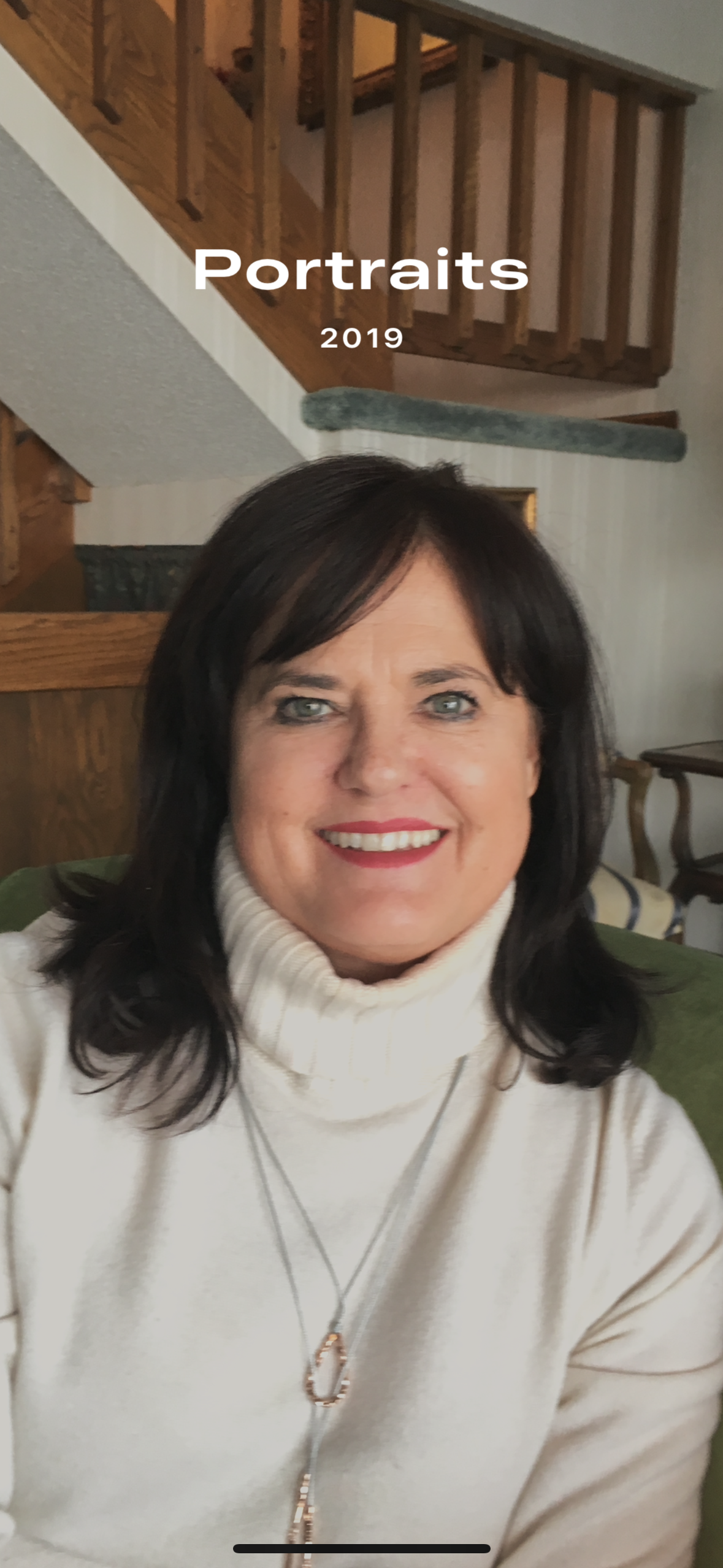 Woman smiling, wearing a white turtleneck and necklace. The background shows a wooden staircase.