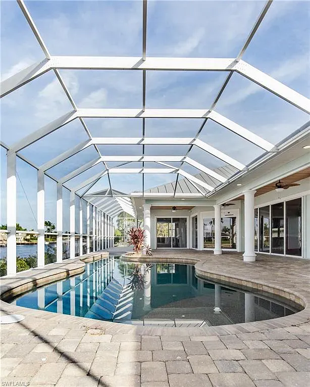 Pool under a screened-in lanai with a view of water, surrounded by a patio and house, blue sky.
