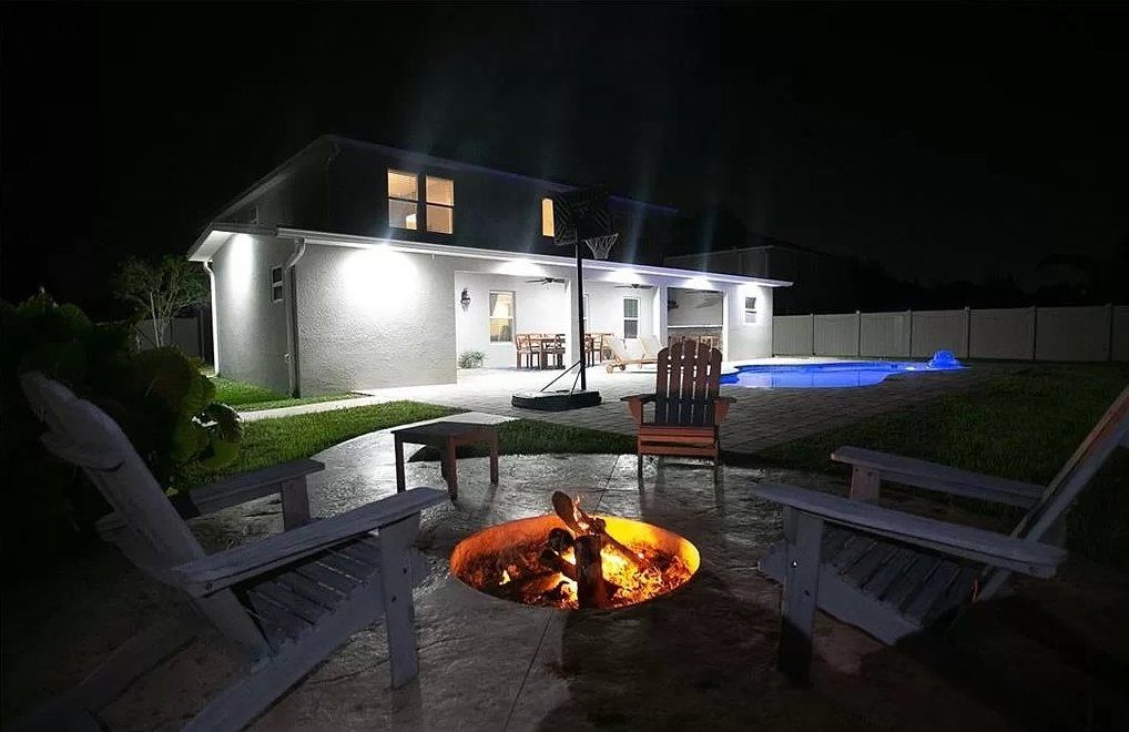 Two Adirondack chairs face a fire pit on a patio at night. A pool and house are in the background.