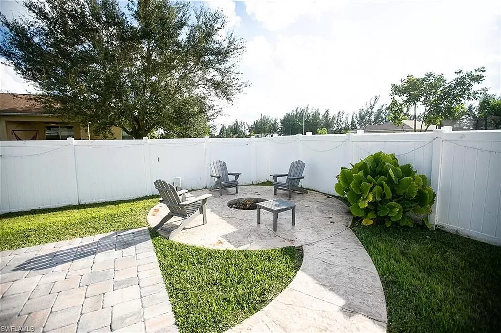 Outdoor patio with Adirondack chairs around a fire pit, enclosed by a white fence and grass.
