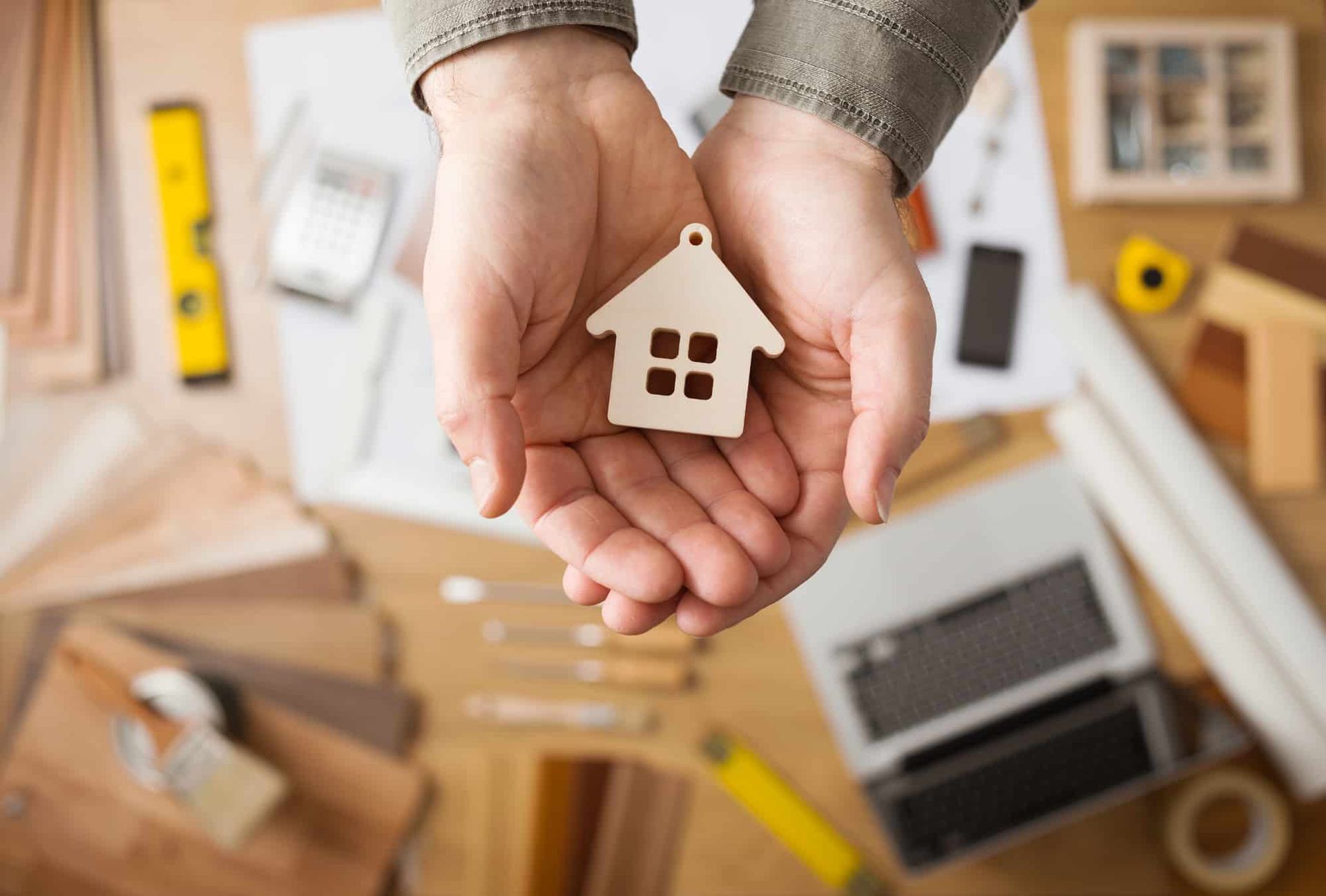 Hands holding a small wooden house figurine; surrounded by tools and blueprints on a desk.