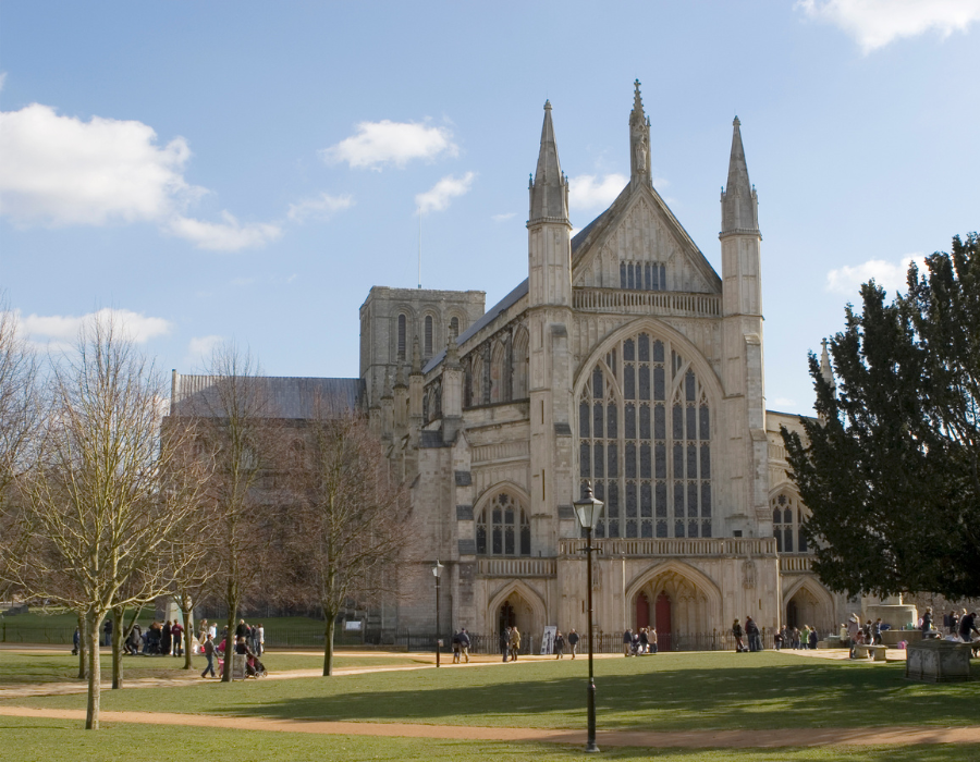 Winchester Cathedral on a sunny day, showcasing its Gothic stone architecture, large stained-glass window, and spires.