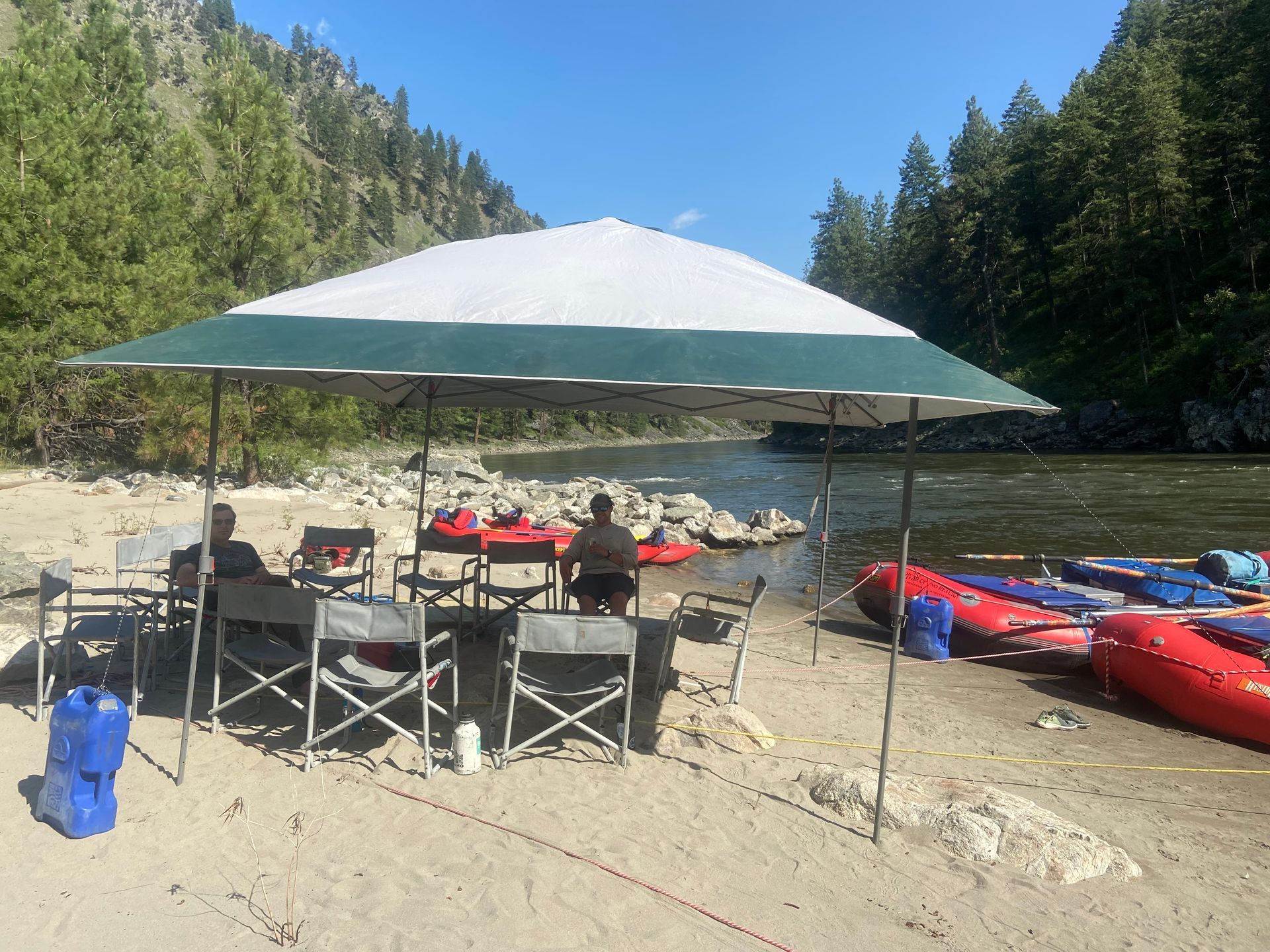 A man is sitting under an umbrella on a beach next to a river.