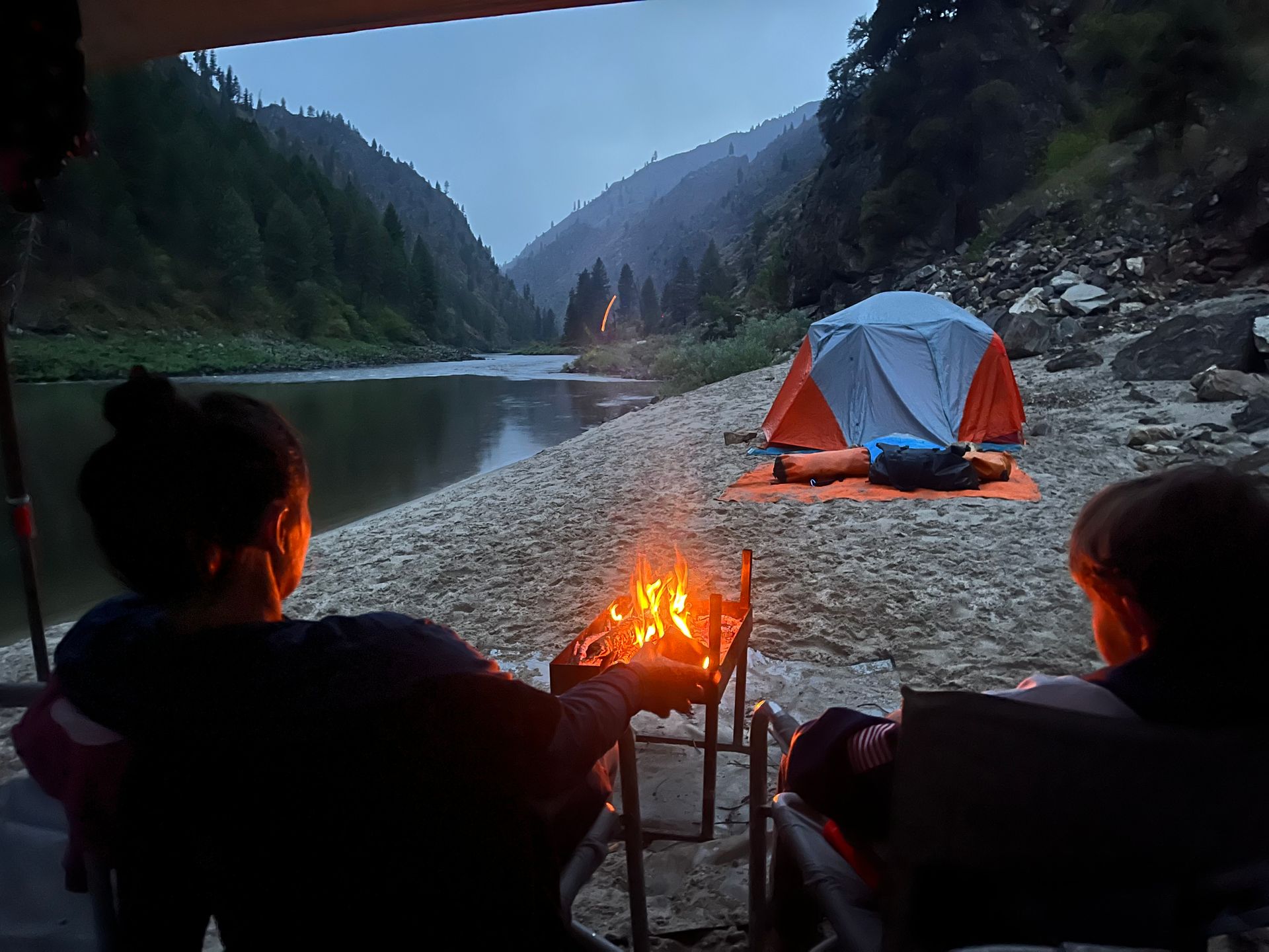 Two people are sitting at a campfire looking at a river and a tent.