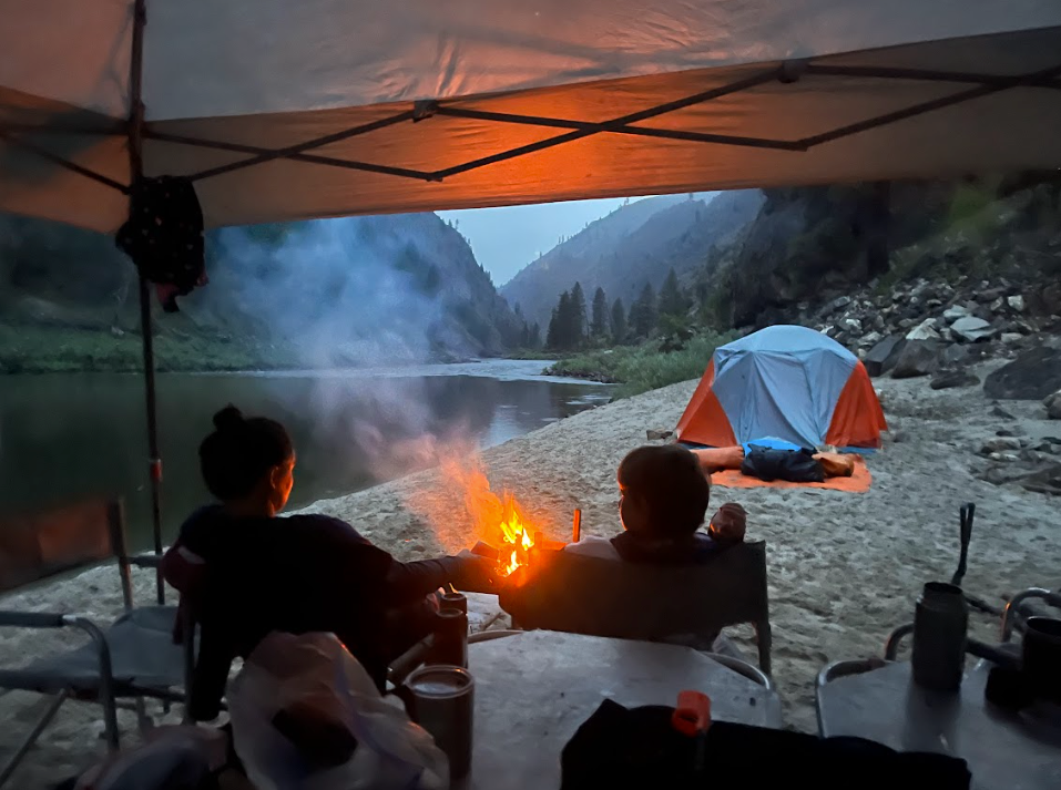 A man and a woman are sitting under a tent looking at a campfire.