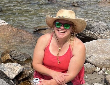 A woman wearing a hat and sunglasses is sitting on rocks near a river.