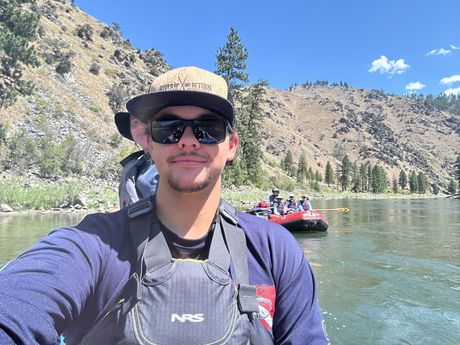 A man wearing sunglasses and a hat is taking a selfie on a river.