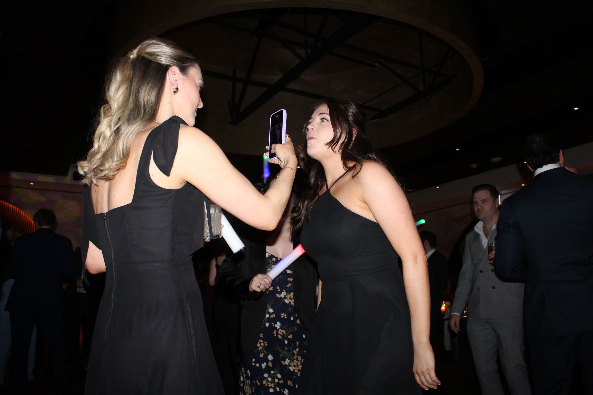 Two women in black dresses dance with glowing light sticks at a party.