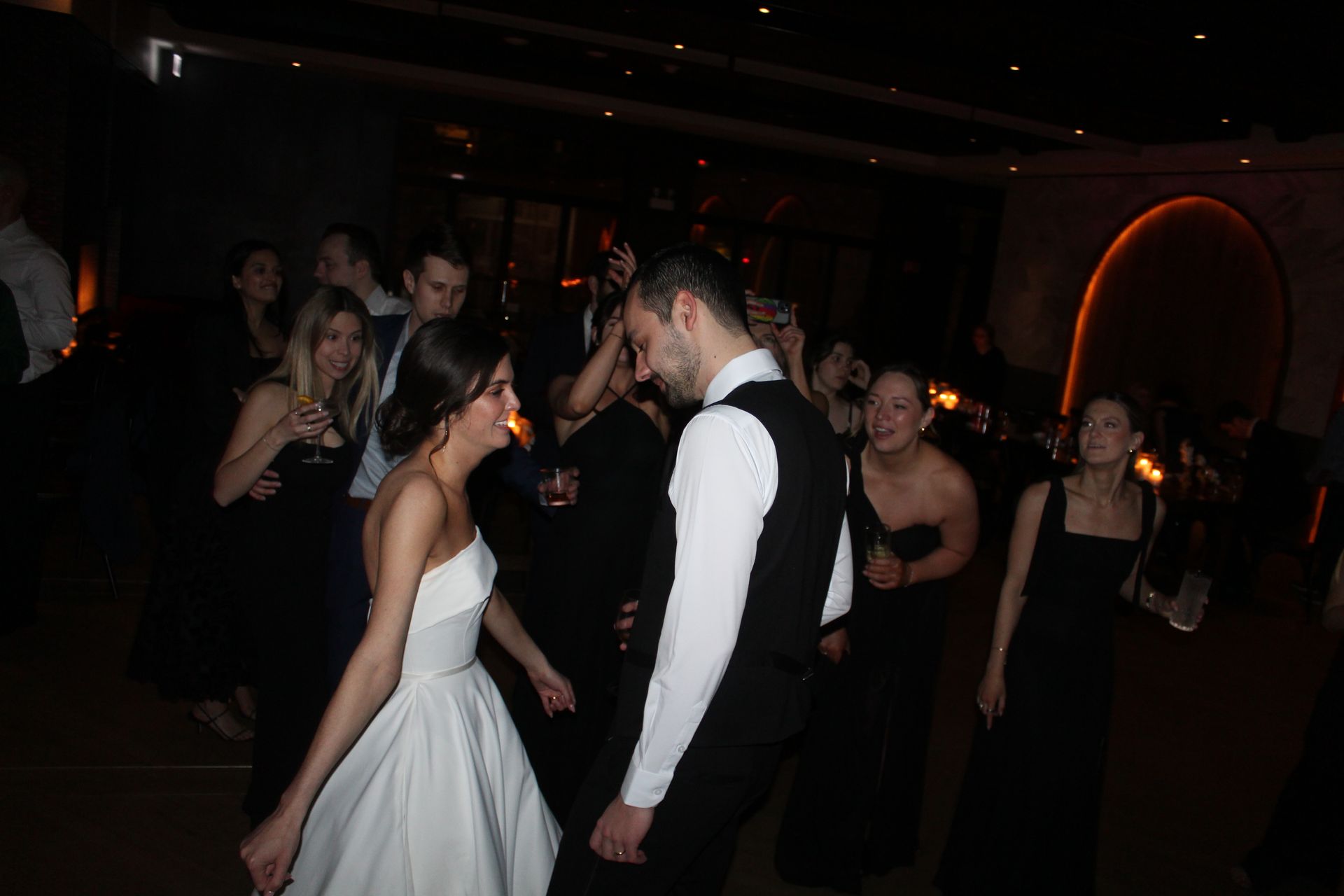 Bride and groom dancing at a wedding reception; others watch and dance, indoor setting, dim lighting.