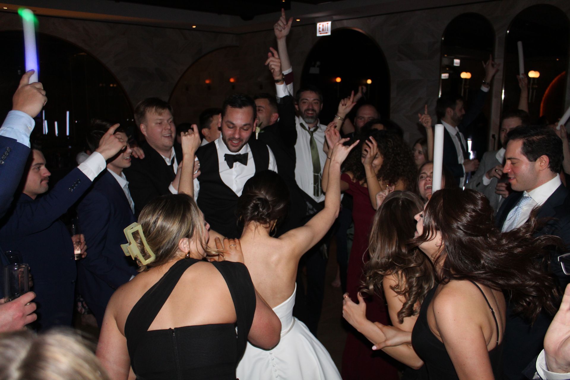 Wedding reception: Bride and groom dance surrounded by guests, some holding glow sticks, arched ceiling.