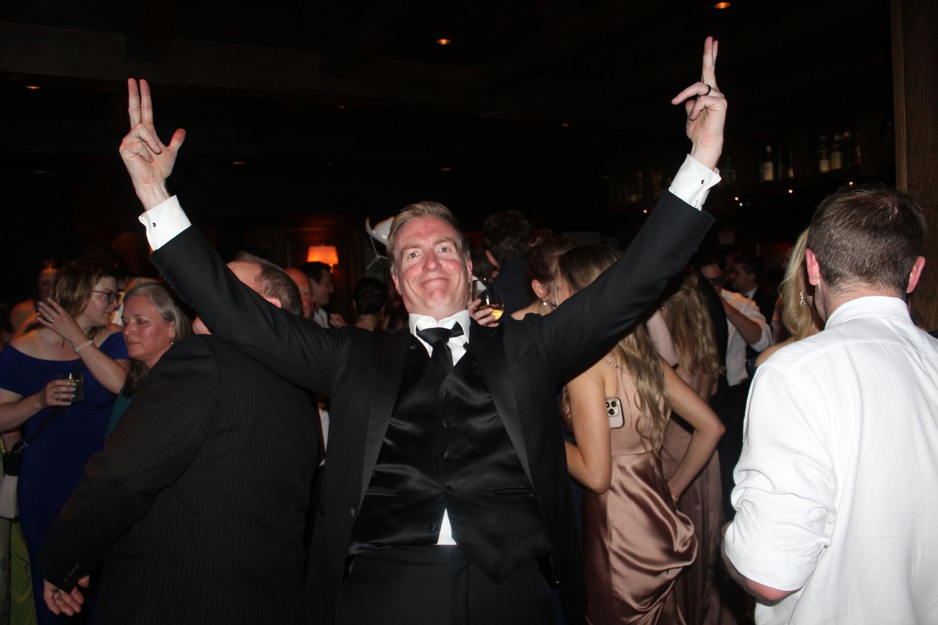Man in tuxedo dances with arms raised and fingers up in a crowded room.