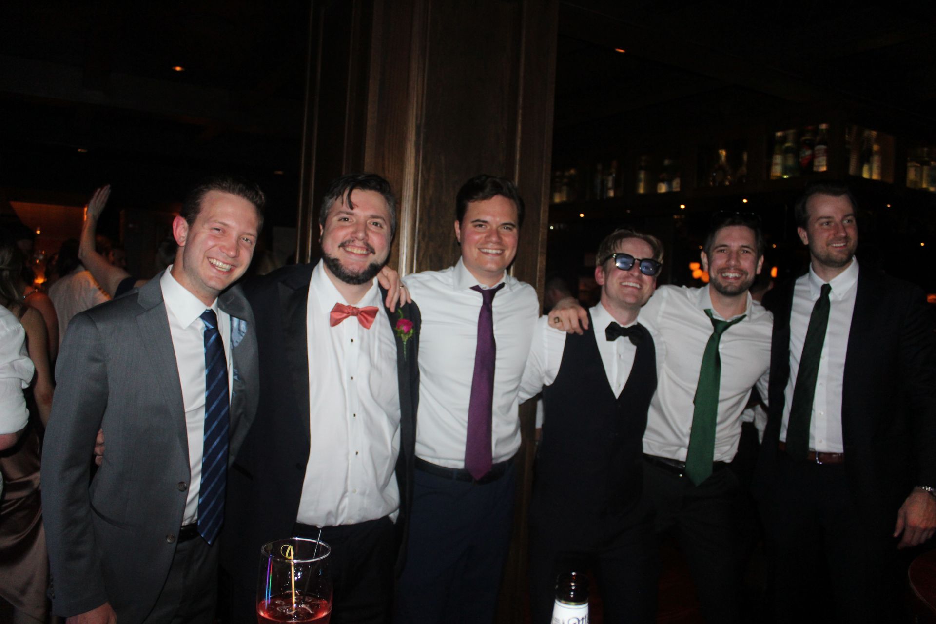 Six men in suits, arms around each other, smiling at a party. Dark interior with gold column.