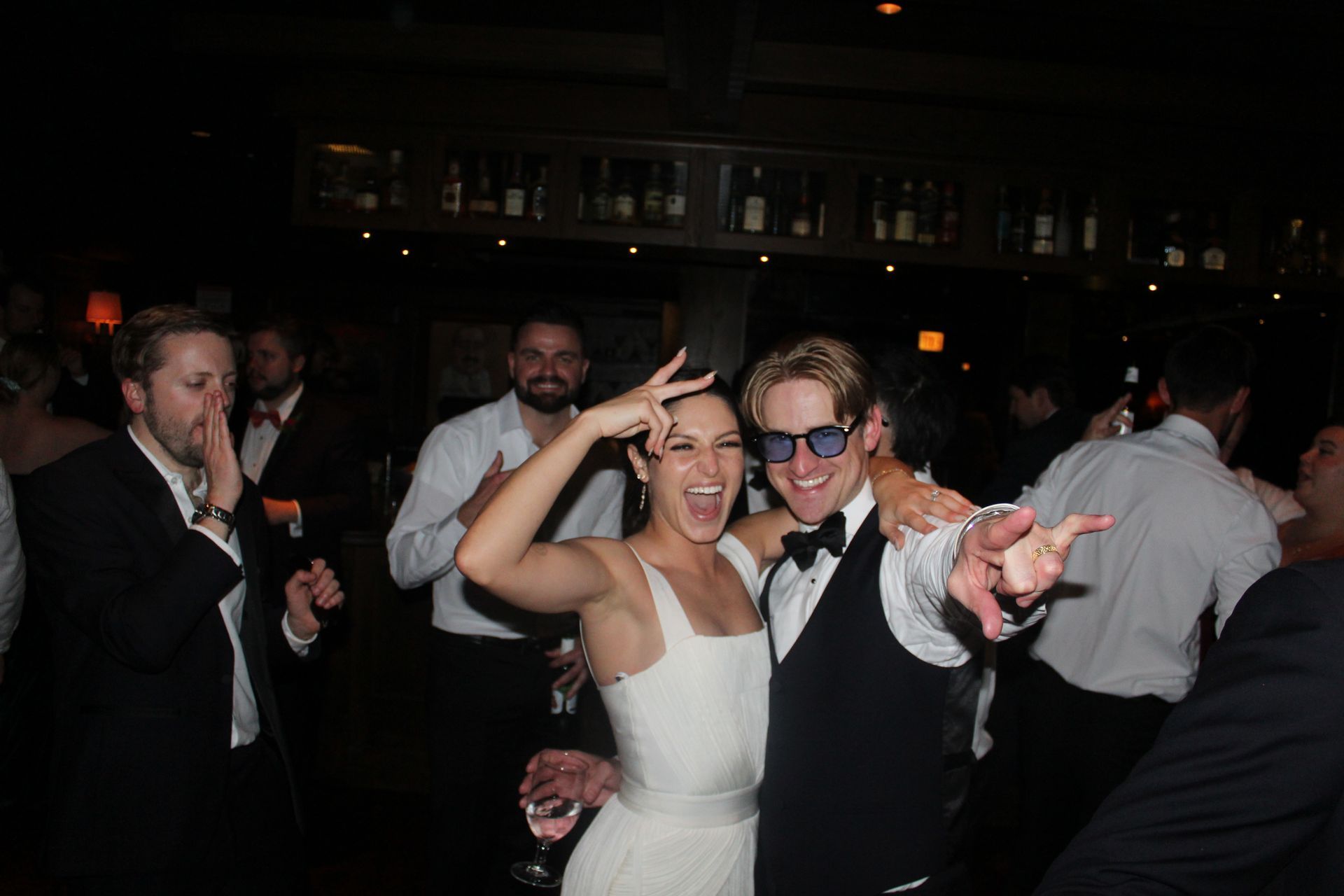 Couple dancing at a party; woman in white dress making peace sign, man in vest and sunglasses points.