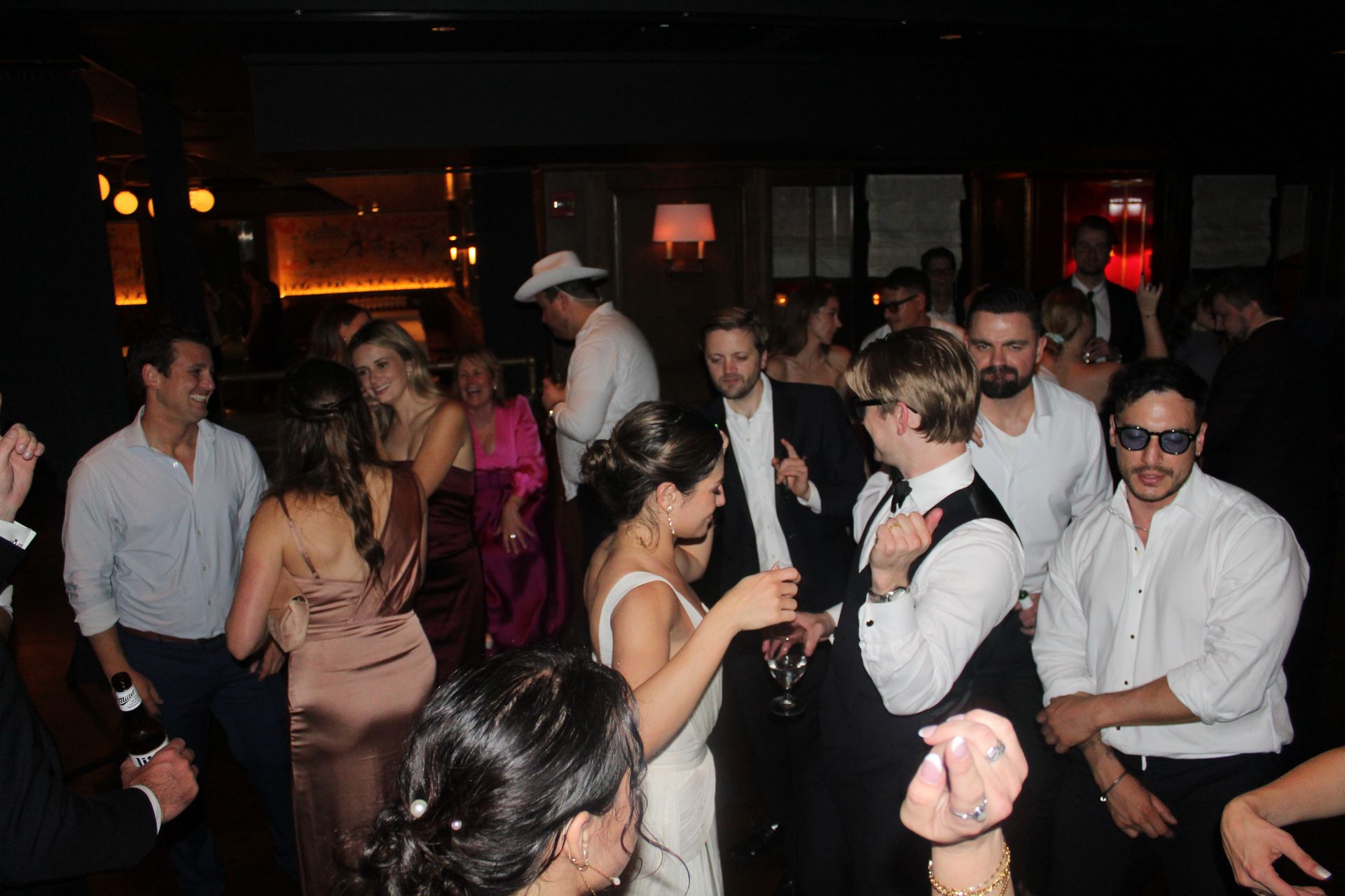 People dancing in a dimly lit club. Several are wearing formal attire, some are holding drinks.