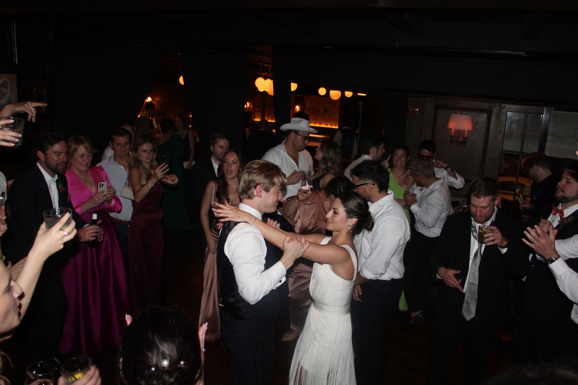 Wedding reception: Couple dancing, surrounded by guests. Dim lighting, formal attire, wooden floor.