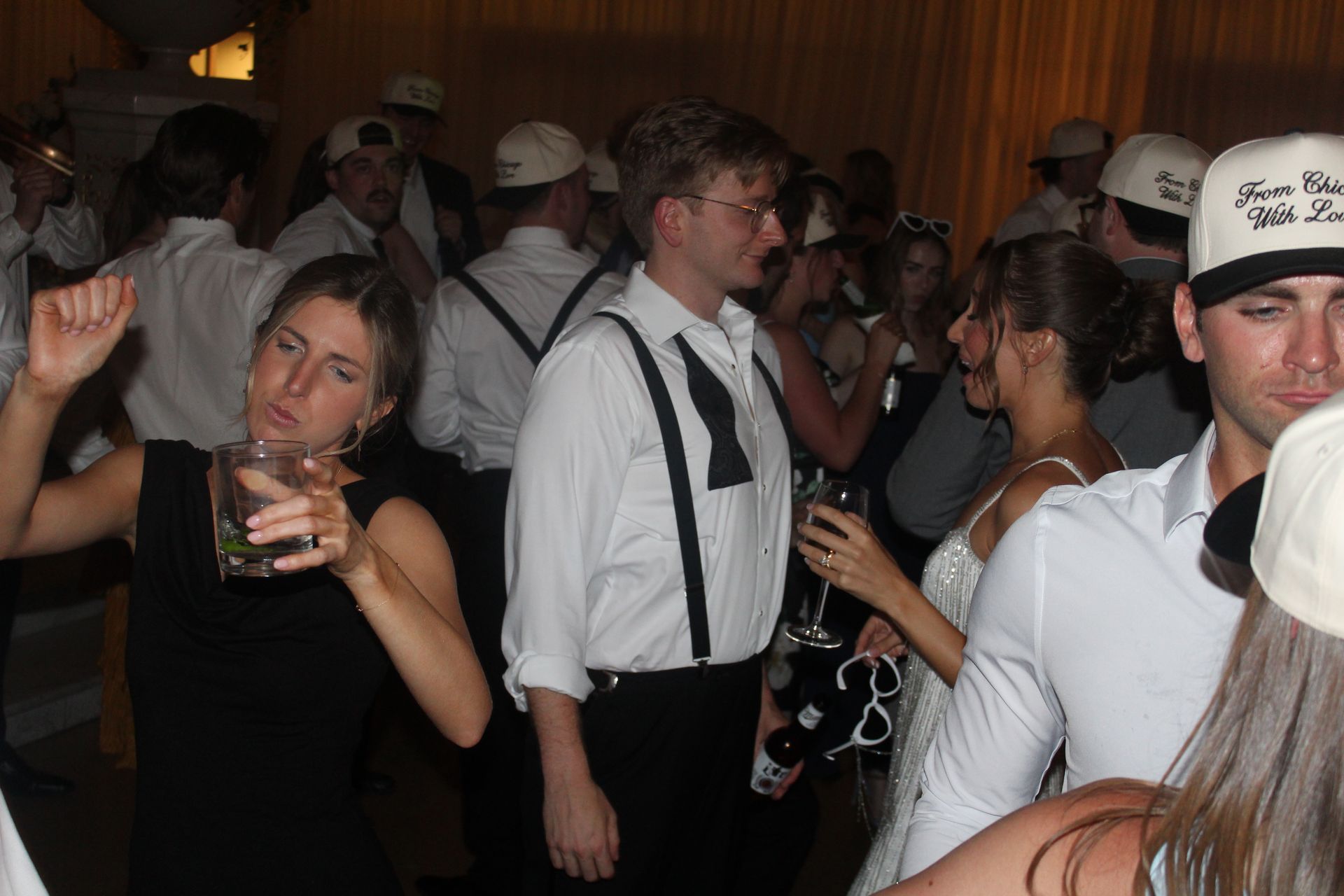 Party scene with people dancing and drinking. Some wear white hats and shirts, a woman in black drinks, others chat.
