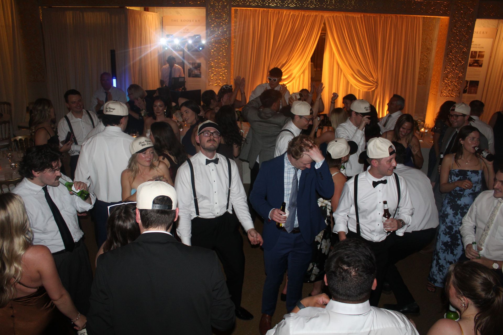 People dancing at an event, many wearing suspenders and hats. Gold curtains in the background.