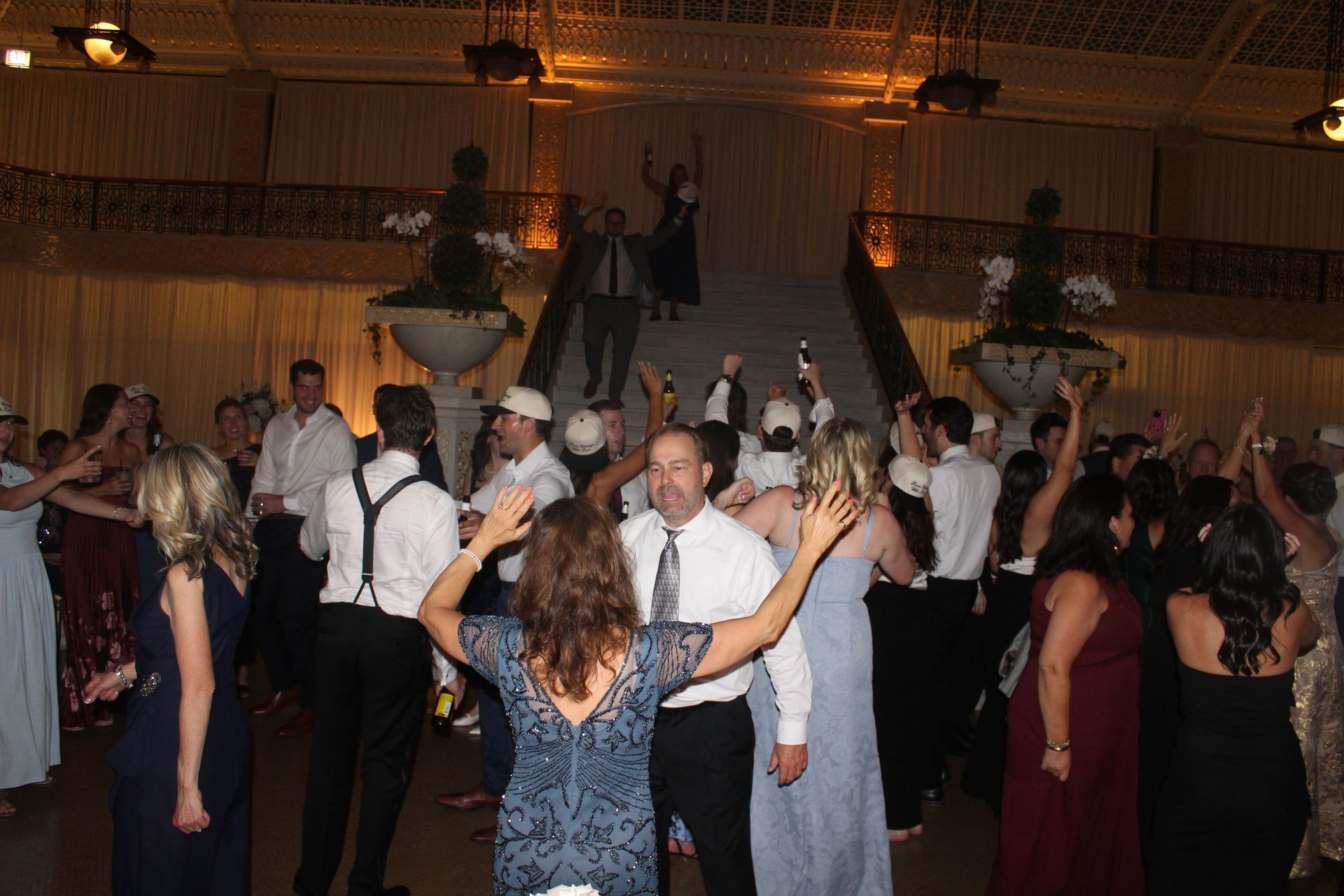 People dancing at a wedding reception; a large ballroom with stairs, some holding drinks, arms raised.