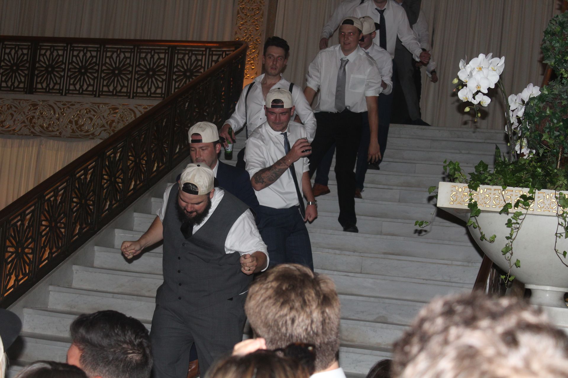 Men in white shirts and baseball caps on stairs, one carried, others walking down. Ornate setting.