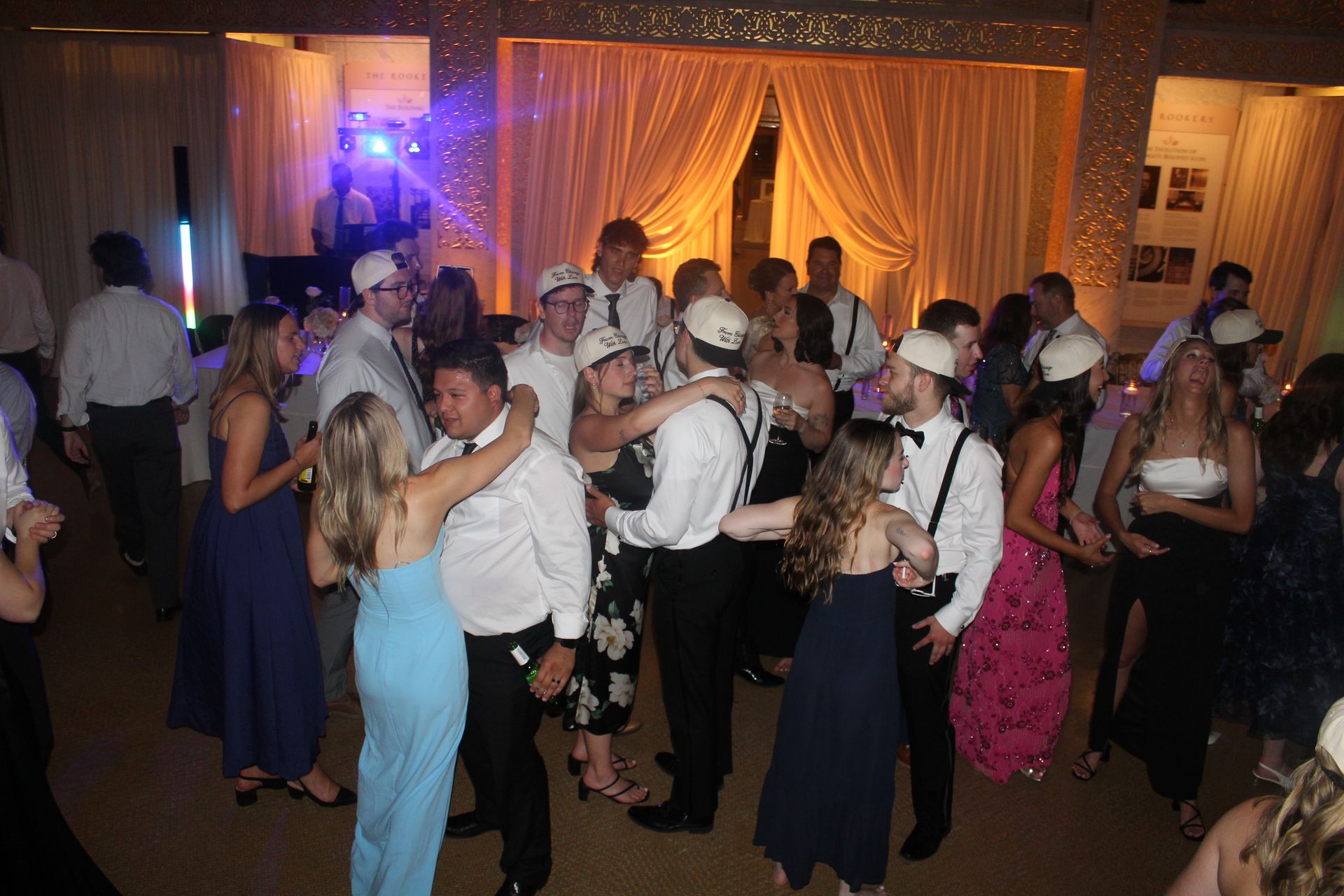 People dancing at a party in a well-lit room with draping and a DJ booth.