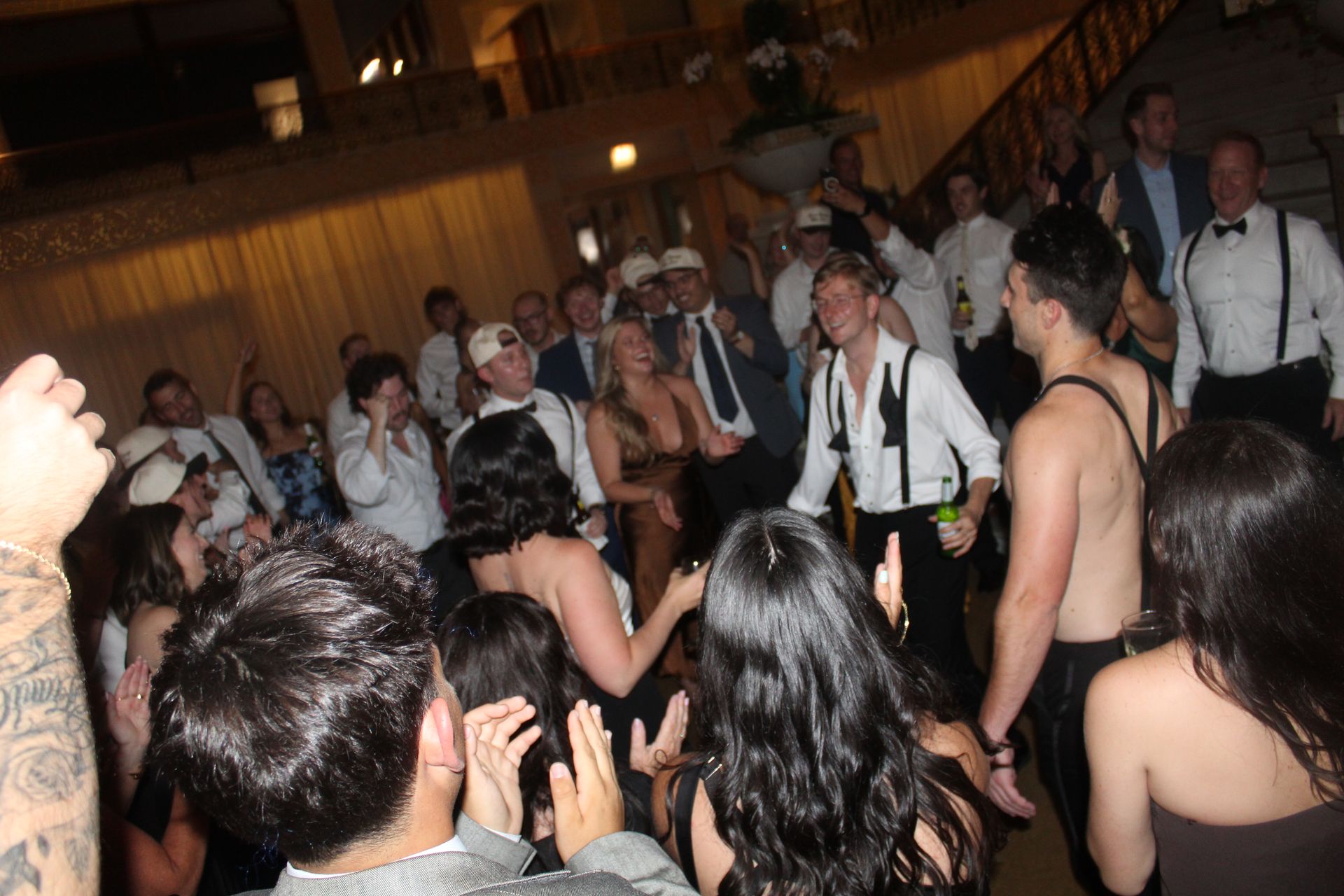 People dancing at an event, some with shirts open. A chandelier and staircase are visible.