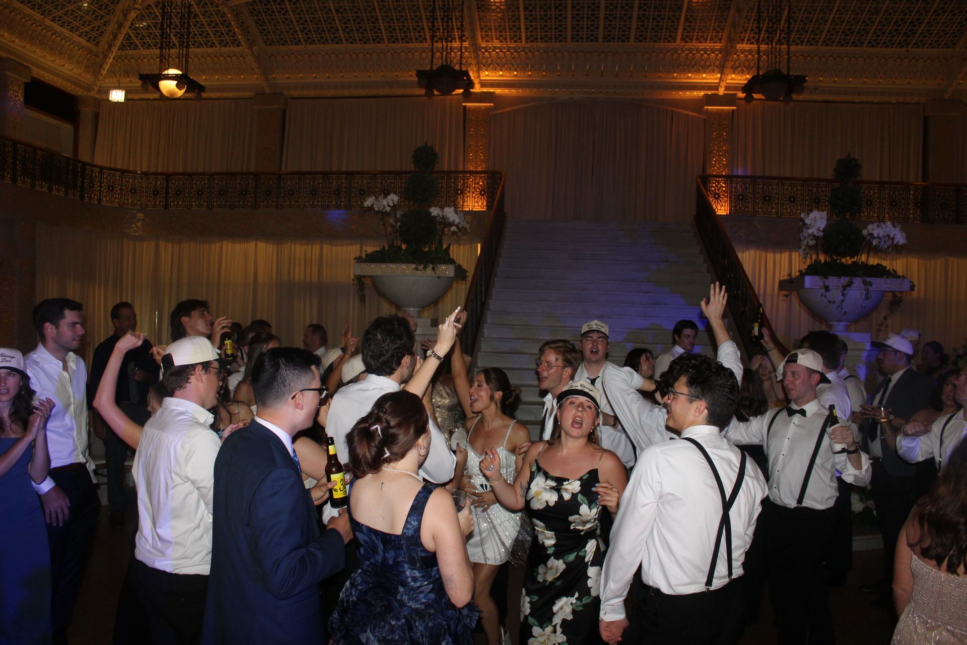People dancing at an event, many wearing white hats. A grand staircase and elegant venue are in the background.